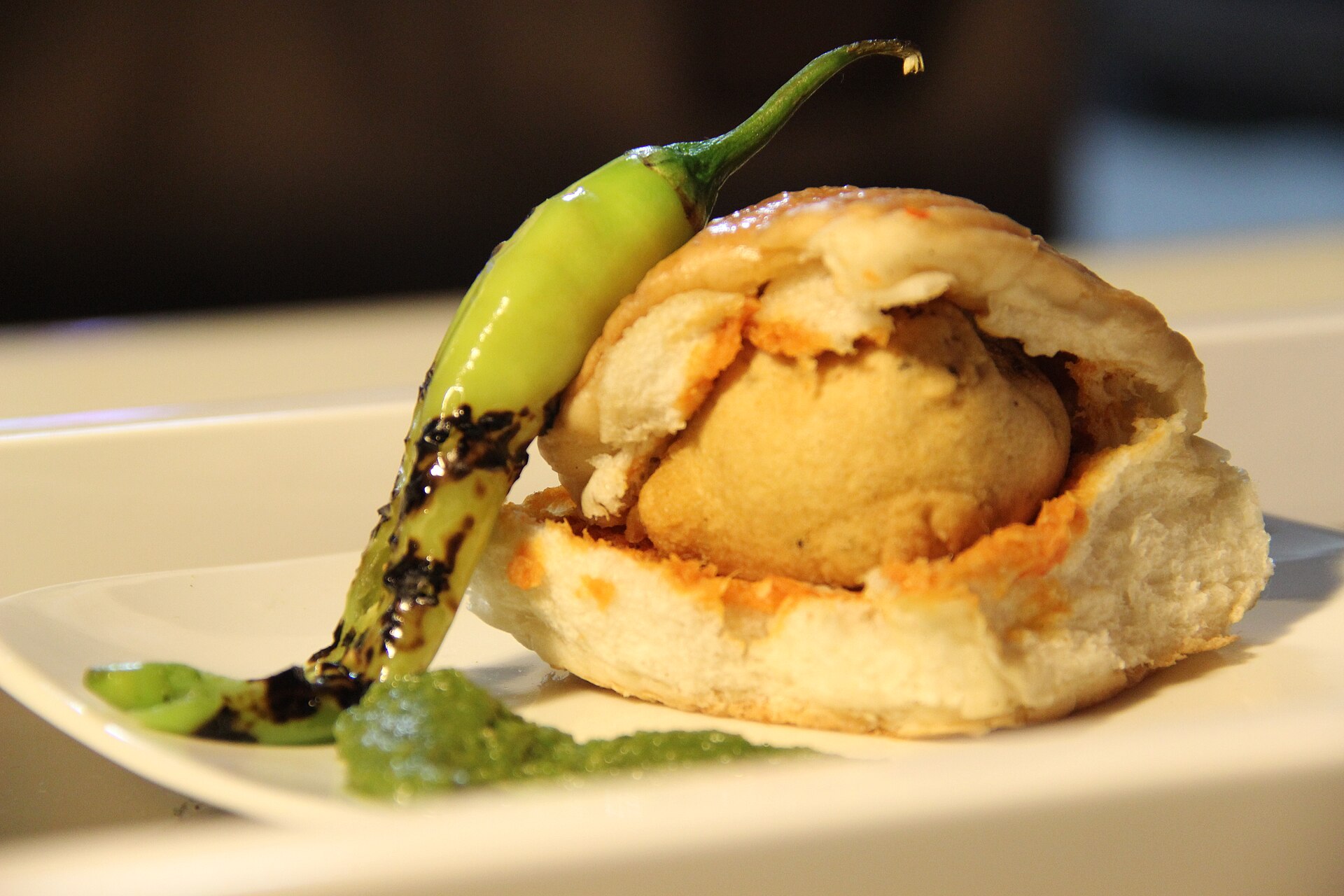 A vada pav with green chutney and a fried chilli on a plate